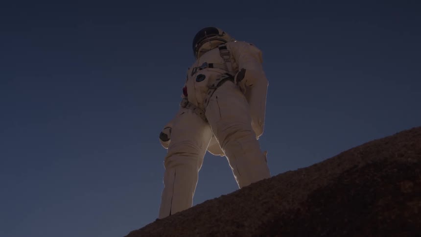 A lowangle view of an astronaut in a white suit standing on a rocky hill under a dark blue sky with a faint glow on the horizon