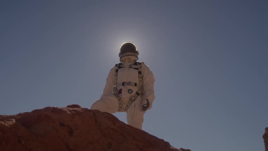 A person in a white astronaut suit with a helmet stands on a rocky hill in a desolate landscape with the sun brightly shining behind their head
