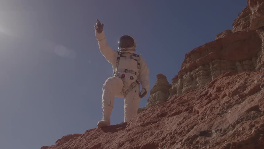 An astronaut in a white spacesuit stands on a red rock formation against a blue sky pointing upwards