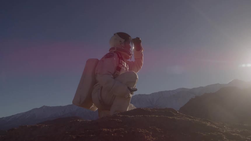 A lowangle shot shows a person in a white spacesuit kneeling on a rocky mound they are facing away from the camera looking out over a mountainous dry landscape under a bright clear sky