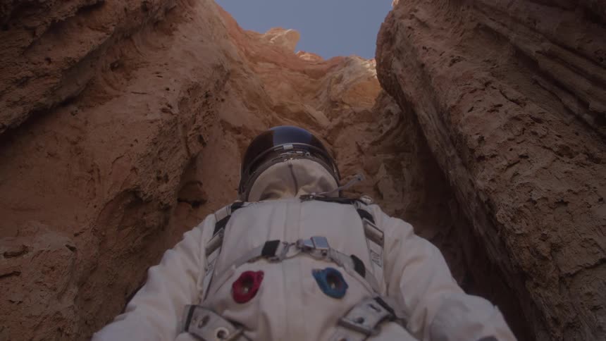 A lowangle view of an astronaut in a white suit with a helmet looking up from the bottom of a narrow canyon with tall rocky walls