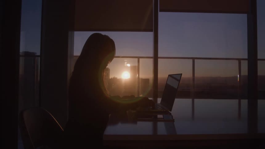 A woman is typing on a laptop keyboard in a office setting at sunset
