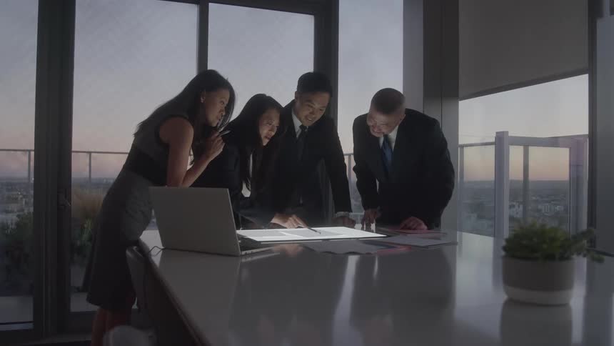 A mediumwide shot shows four business professionals gathered around a conference table in an office with large windows they are all leaning forward looking down at a brightly lit document on the table
