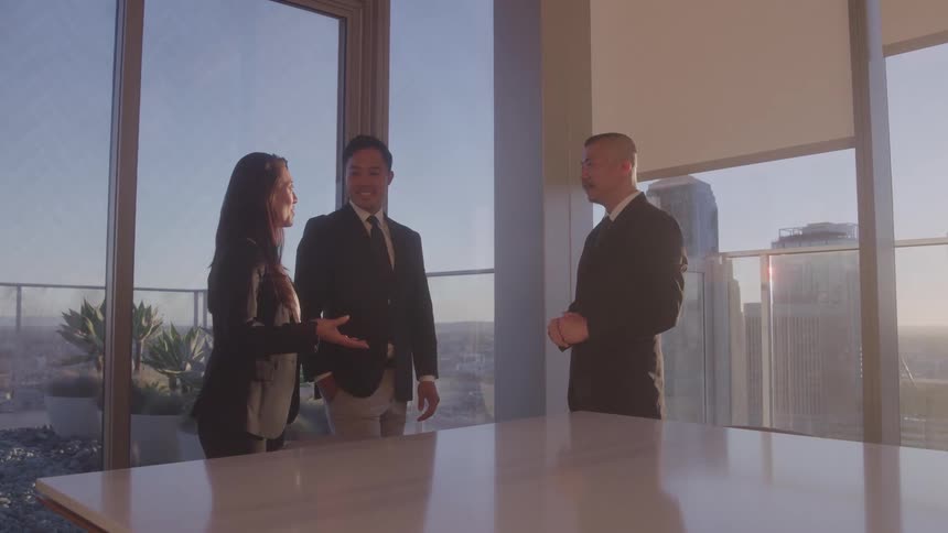 Two men and a woman all wearing suits standing in a highrise conference room at midday engaged in a business discussion