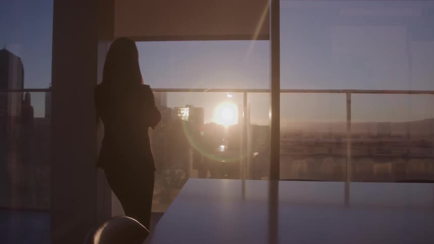 A silhouette of a person stands near a window looking out over a city at sunset the bright sun casts a golden glow and lens flares into the room