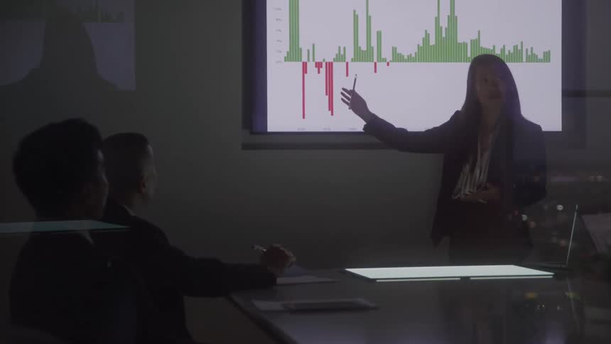 A woman is presenting a financial chart to two male colleagues in a dark room with a glowing table and computer