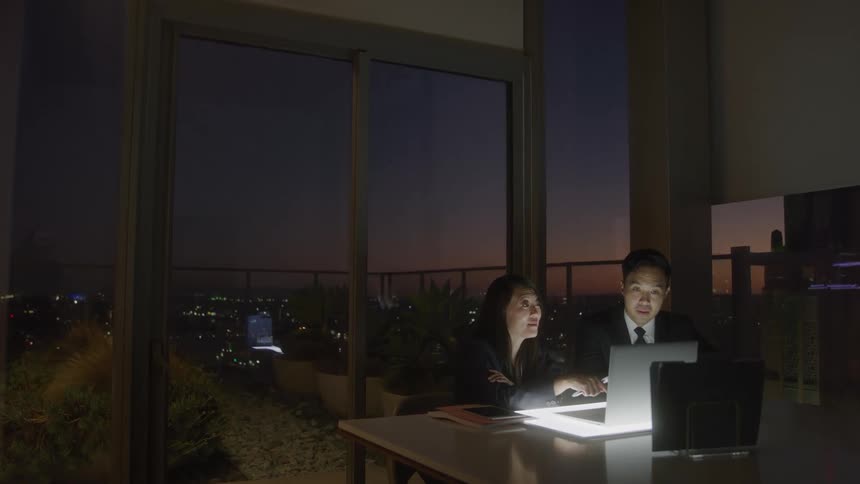 Woman and man sitting behind a desk in an office at night with a laptop in front of them as the woman explains details of the business displayed on the screen