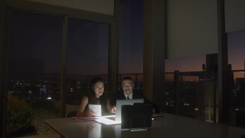 Two people in an office at night sit at a desk with a laptop and paper and talk