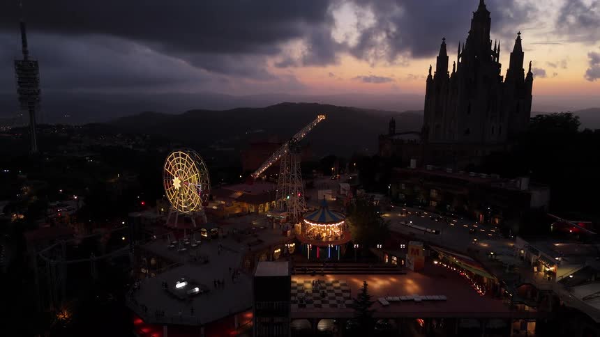 Amusement park rides operate at a theme park overlooking a valley while a red afterglow tints the horizon