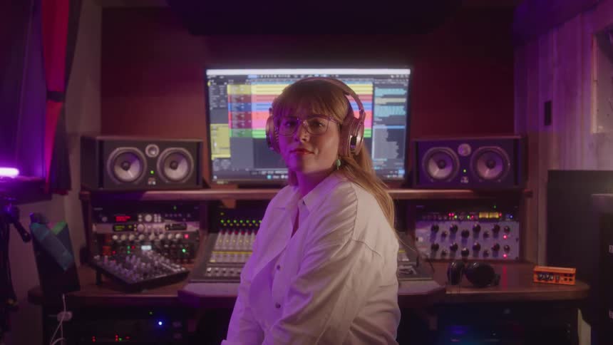 Person wearing headphones and glasses sitting in a recording studio looking at the camera with an audio mixing screen in the background