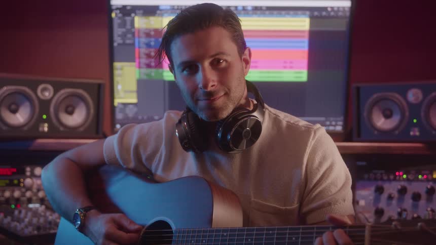 A man sitting at an audio editing desk wearing headphones around his neck with music editing software opened on the monitor behind him playing acoustic guitar and smiling