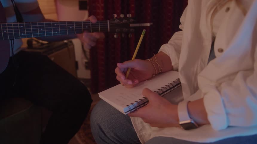 A close view of a person with a pencil and a notebook sitting in front of a man playing a guitar with a lamp and a tapestry in the background