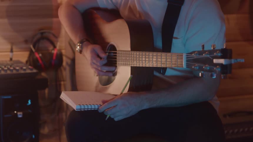 A person strums an acoustic guitar while jotting in a notebook appearing thoughtful and creatively absorbed