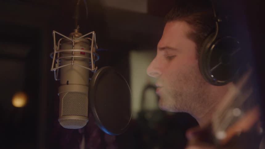 A close view shows a man with a beard and headphones on his head singing into a large condenser microphone in a dimly lit recording studio