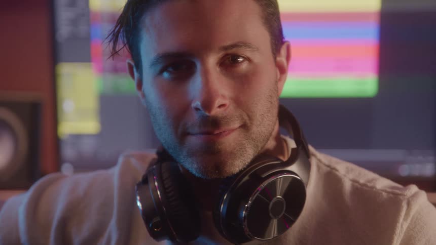A man with headphones around his neck smiling with his back to a computer station with music editing software opened on the monitor