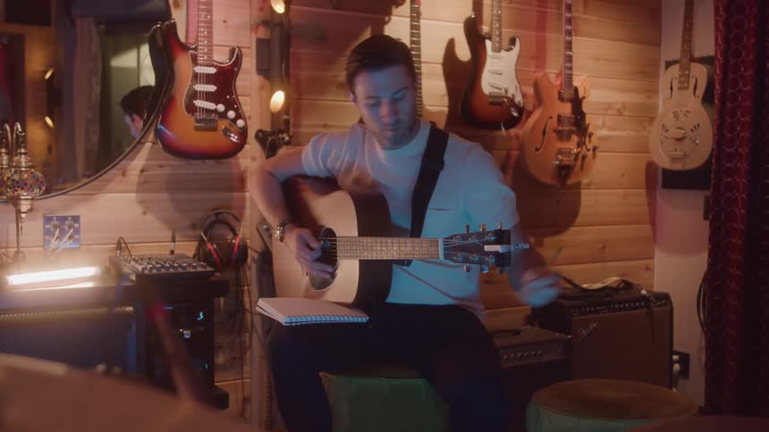 A man sits in a cozy woodpaneled music room intently strumming an acoustic guitar with a notebook on his knee while several electric guitars hang on the wall