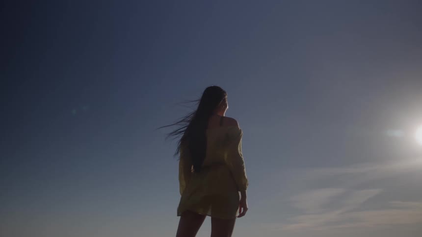 A long view shows the silhouette of a woman with long hair and a lightcolored dress standing outside with her back to the camera against a clear bright sky