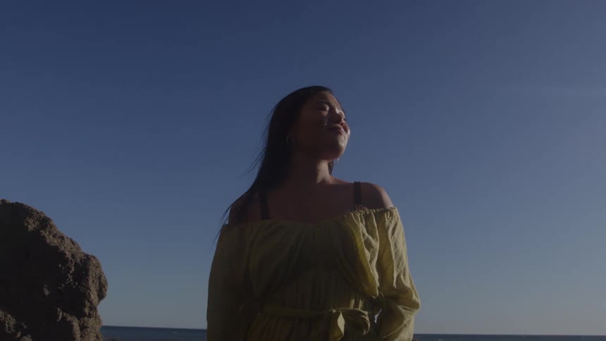 A woman stands by the sea with her face lifted toward the sun eyes closed and smiling in peaceful contentment