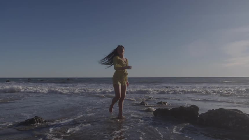 A woman walks toward the ocean hair loose and arms lifted looking free and carefree