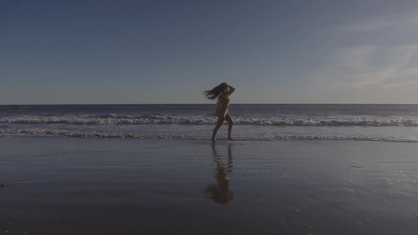 A silhouette of a woman in a lightcolored dress and with long hair walks along a wet beach with gentle waves and a bright sunset on the horizon