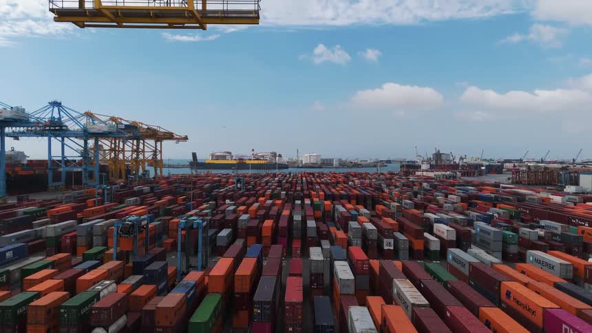 A drone shot of a commercial port with hundreds of colorful shipping containers stacked in neat rows with large yellow cranes and a cargo ship visible in the background under a partly cloudy sky