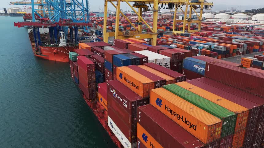 A drone shot of a large cargo ship being unloaded by a giant crane at a commercial port with hundreds of colorful containers stacked high