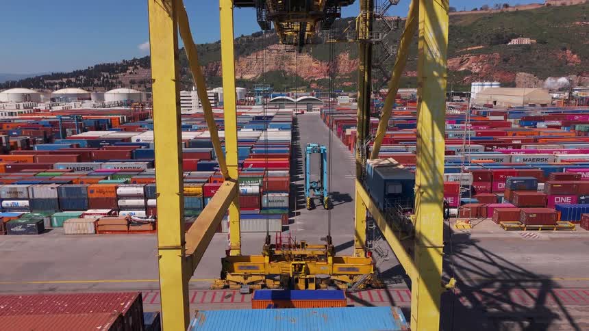 Men work with loading cranes and mobile loaders at a loading dock located near a mountain on a sunny day