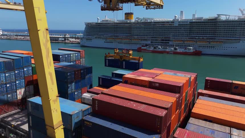 Based on the video a large crane is moving a blue shipping container and placing it on top of a stack of containers on a cargo ship in the background there is a large white cruise ship docked at the port