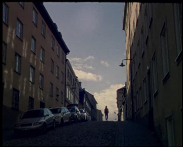 Man walking along the narrow road between apartment buildings passes by a row of parked cars on a cloudy day
