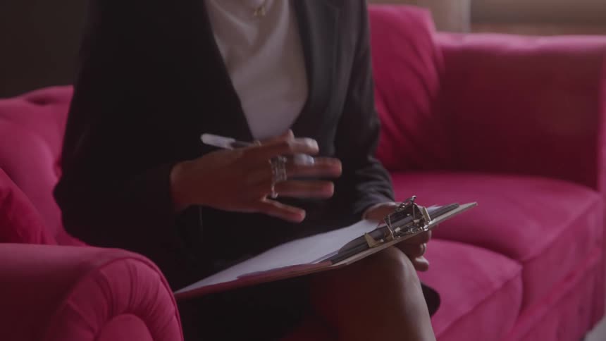 A close view shows a woman wearing a black jacket sitting on a plush red couch and taking notes on a clipboard with a pen