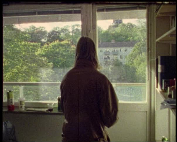 A man with his back to the camera walks out a door to a sunlit suburban neighborhood