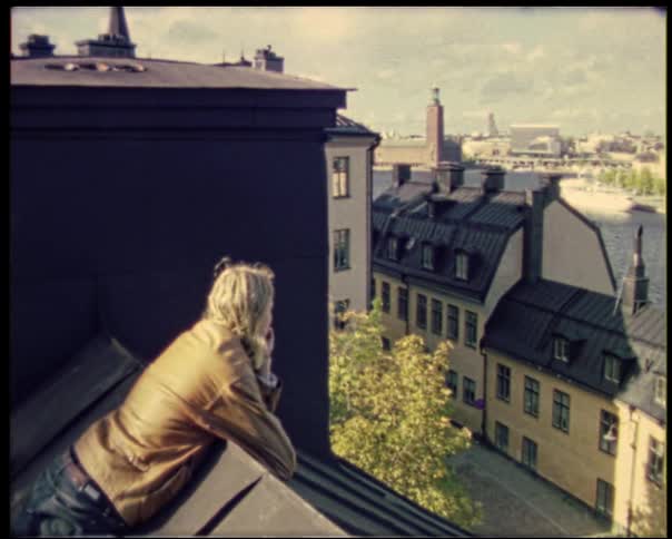 Man in a brown jacket leaning on the rooftop overlooking the city