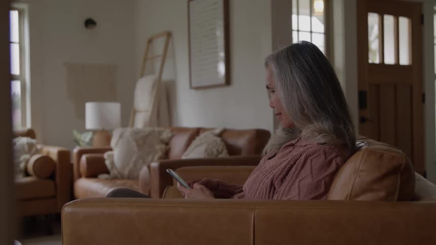 A woman sitting alone on a one person couch in a living room using an app on her smartphone