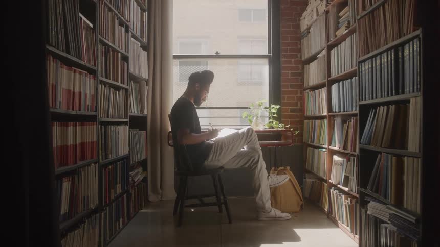 Based on the video a man is sitting on a chair in a room with floortoceiling bookshelves he is looking down and writing in a notebook