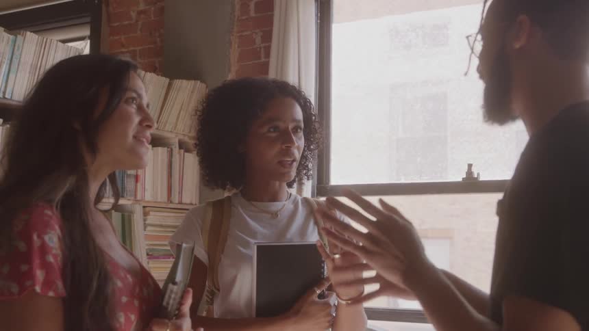 A young man with glasses and a beard is standing near a window and talking with two young women in a room with a bookshelf in the background