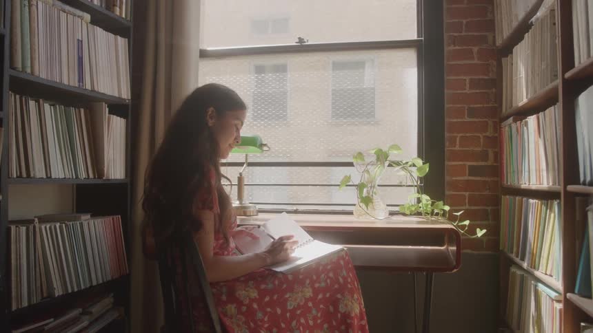 A woman sitting by a small table next to a window with bookshelves on both sides of her filled with notebooks writing on a large notebook and smiling