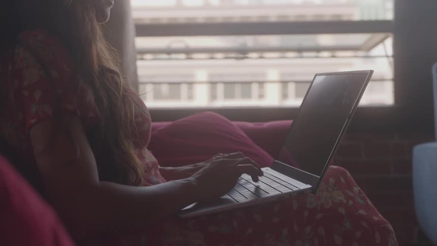 A woman with dress is typing at a laptop sitting on a sofa in a home office setting