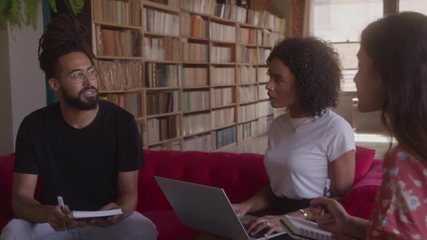 Three diverse colleagues collaborate on a project sitting on a red couch in a book lined room while using a laptop and notebooks