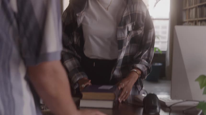 A smiling woman with curly hair is standing in a room with a wall full of bookshelves she picks up a book and places it on a table in front of her then looks at a laptop