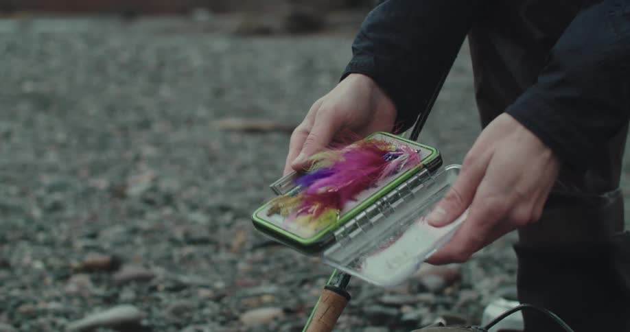 A person opens a small tackle box to reveal colorful fly fishing lures