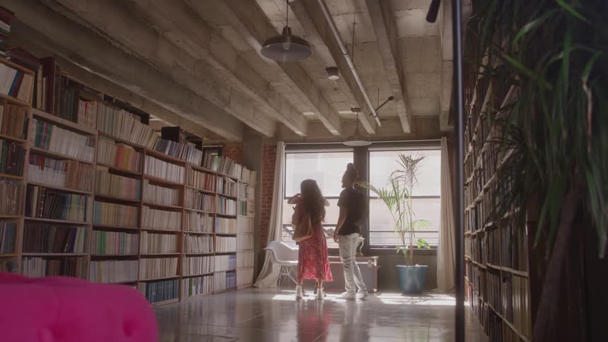 A man and woman walk into a modern library passing a woman sitting on a trunk who is reading they stop and talk in front of a window the room is filled with bookshelves and has concrete ceilings with exposed beams