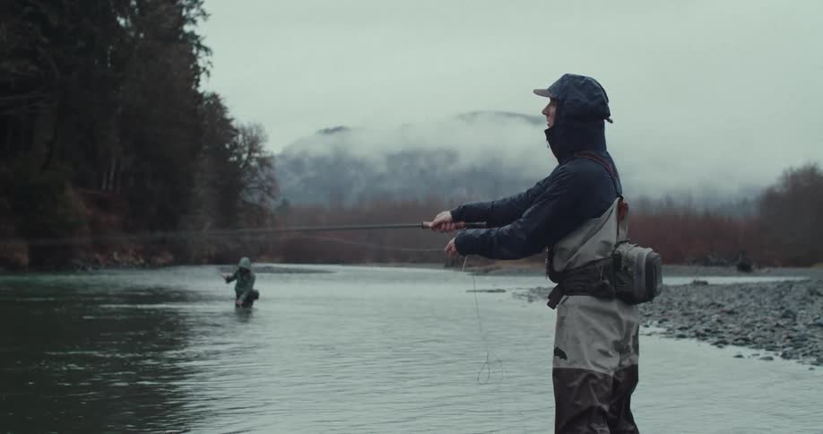 A man wearing waders and a black jacket is fly fishing in a river with another person fishing in the background and a lush treelined bank under a cloudy sky