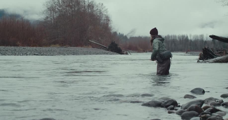 A woman standing in a river with the water up to her knees using a fishing pole to throw a fishing line