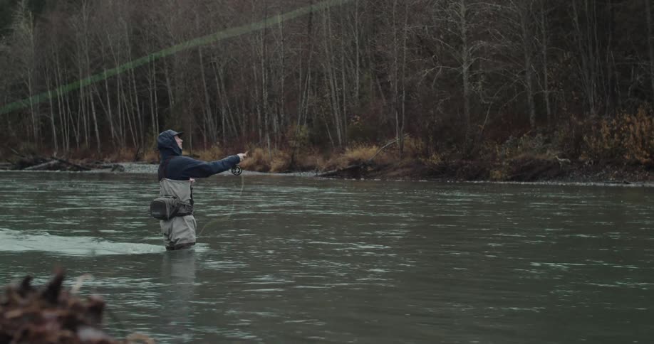 A person in a hoodie and waders is standing in a river casting a fishing line the river is surrounded by a forest of bare trees and the sky is overcast