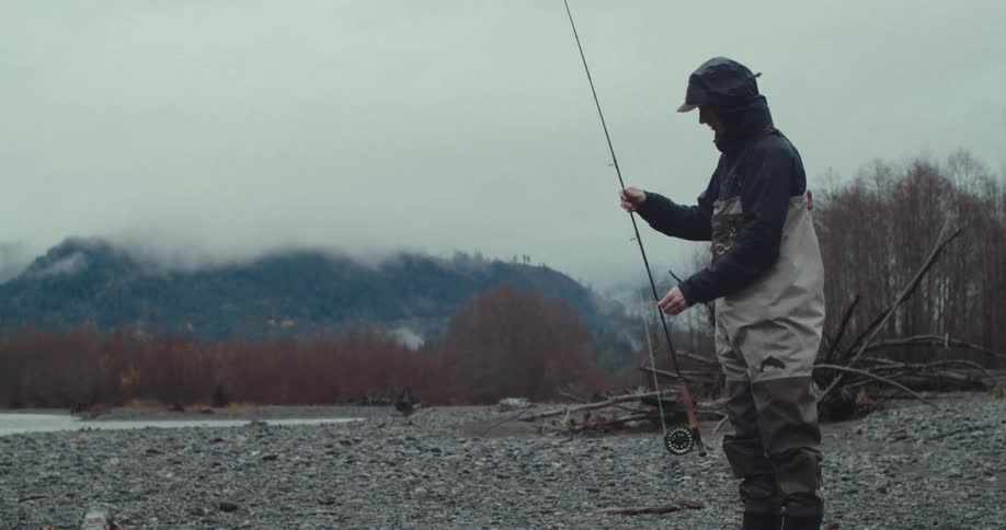 Young man standing on a rocky riverbed setting up his fishing line dressed in synthetic base layers and quickdrying pants