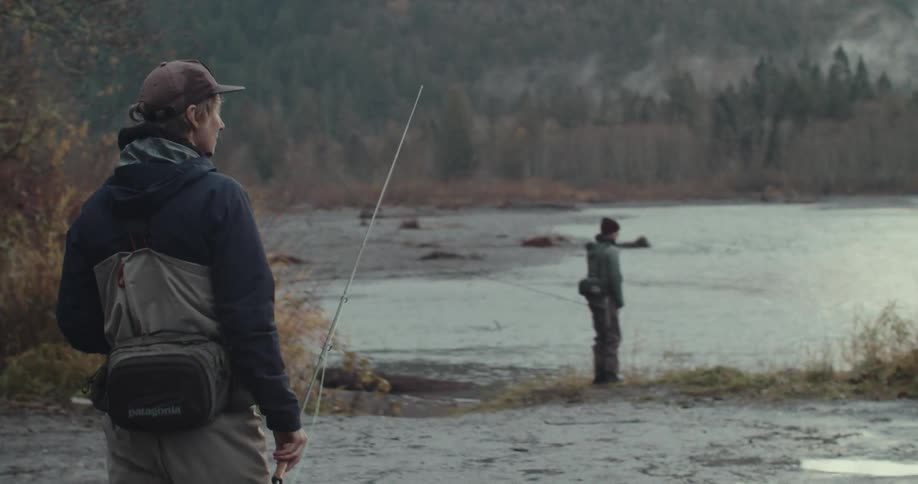 A fisherman in waders walks towards a river where another person is fishing
