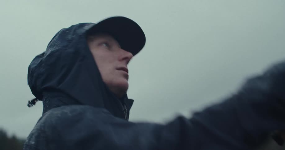 A closeup shot shows a person wearing a dark jacket with a hood and a baseball cap underneath the person looks focused and rain is visible on the jacket the background is blurry with a grey sky and distant trees