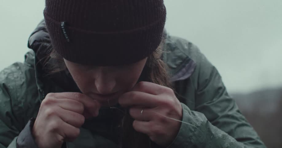 A closeup shot shows a person wearing a beanie and a green jacket the person is focused on their hands tying a knot in a thin clear fishing line the background is a blurry overcast sky
