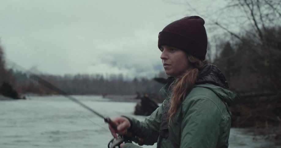 A person wearing a beanie and a green jacket is fishing on a riverbank the person is holding a fishing rod and looking off into the distance the background is a grey sky and a river with trees on the other side