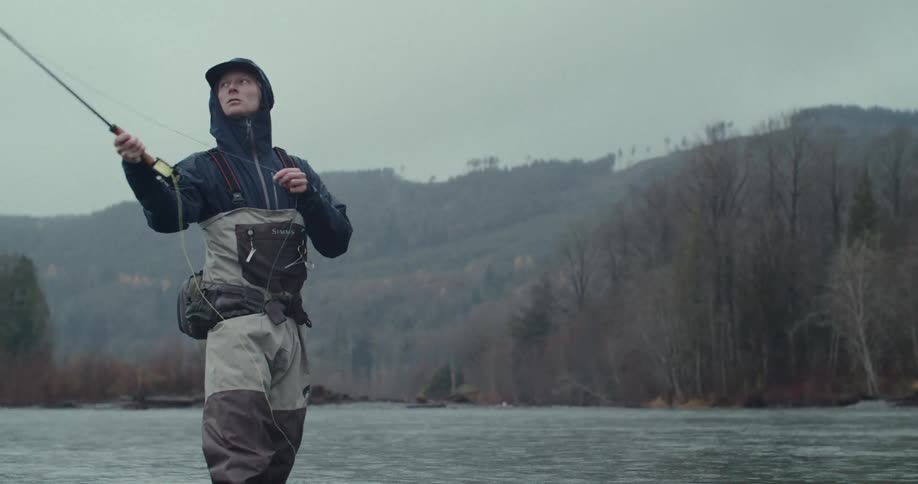 A person wearing waders and a rain jacket is fly fishing in a river the person is focused on their line which extends out over the water the background is a mountain covered in bare trees under a grey sky